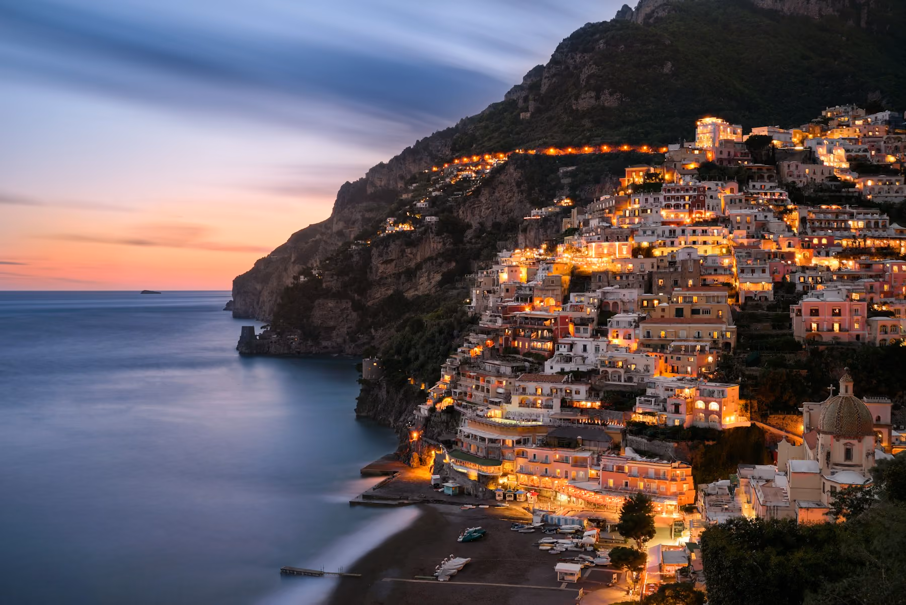 Positano lit up at dusk above a calm sea: the best time to visit Amalfi Coast towns is May, June or September