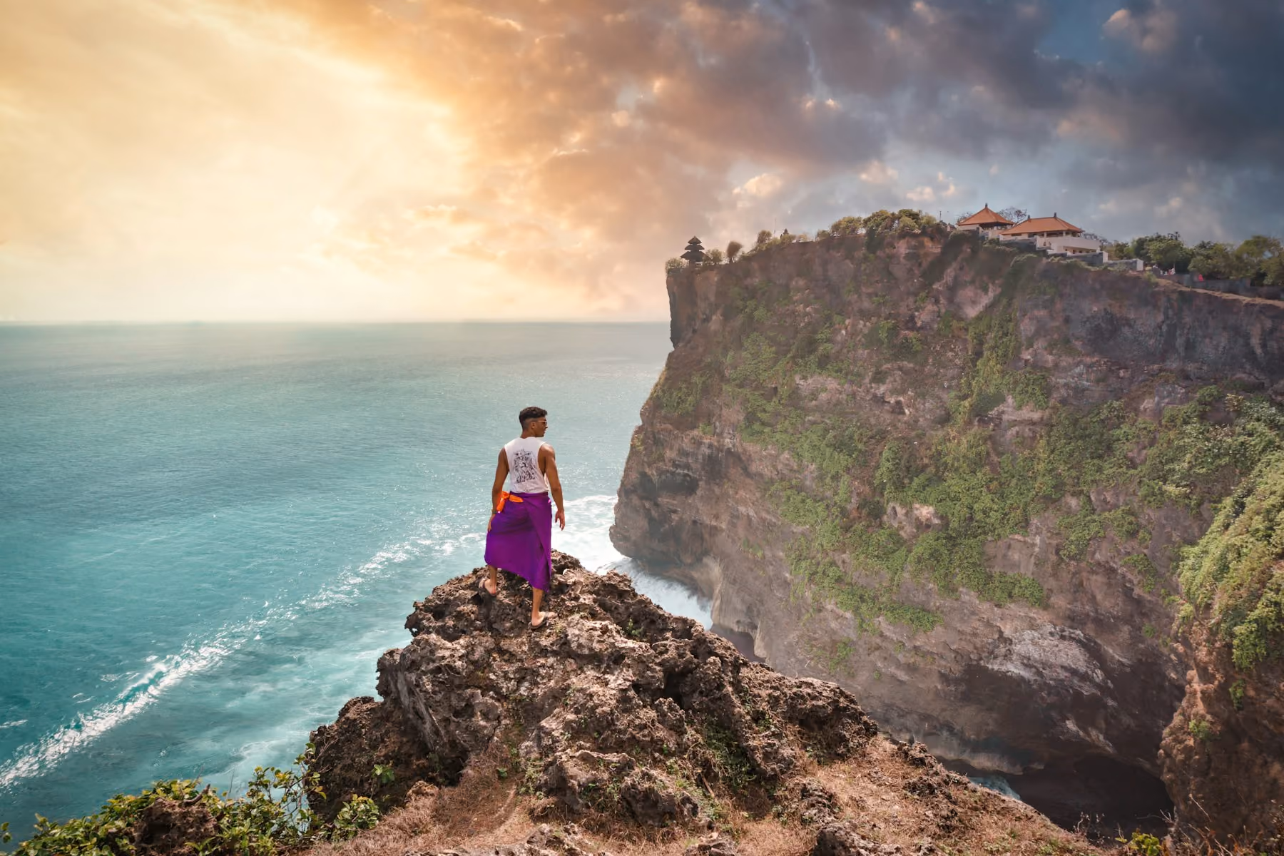 A visitor on a rocky outcrop above turquoise water, with Uluwatu Temple on the cliff edge at sunset