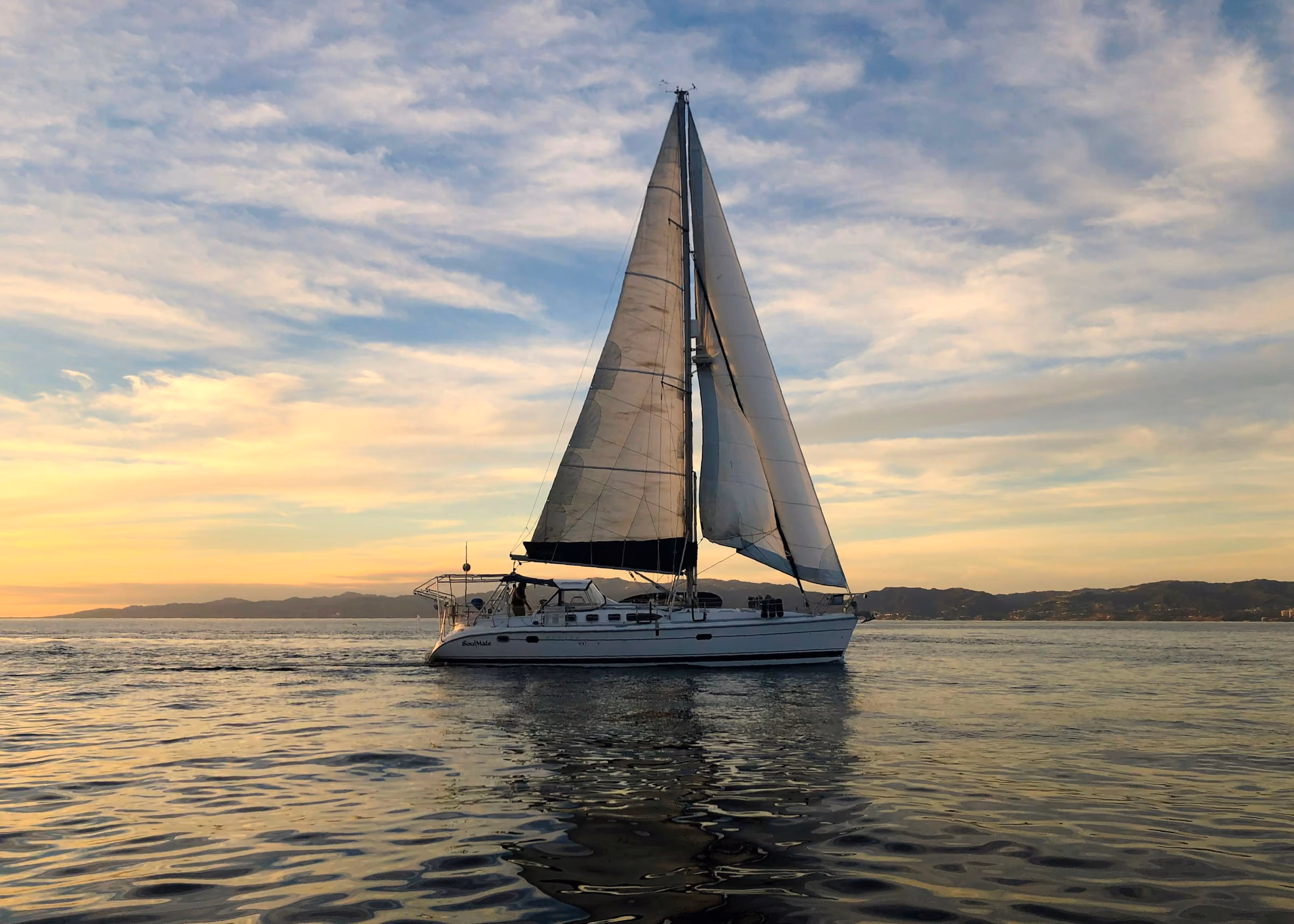 A sailing catamaran under full sail at sunset, the sea flat and gold