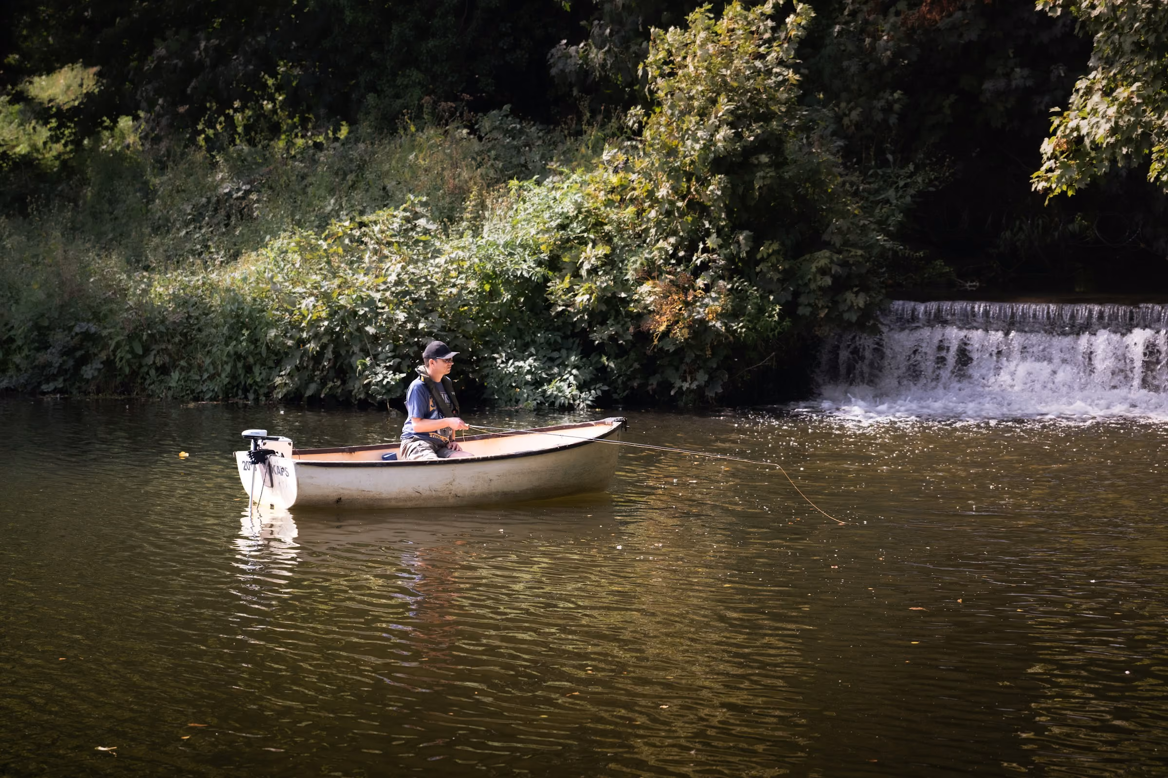 An angler in a small boat below a weir on a wooded river