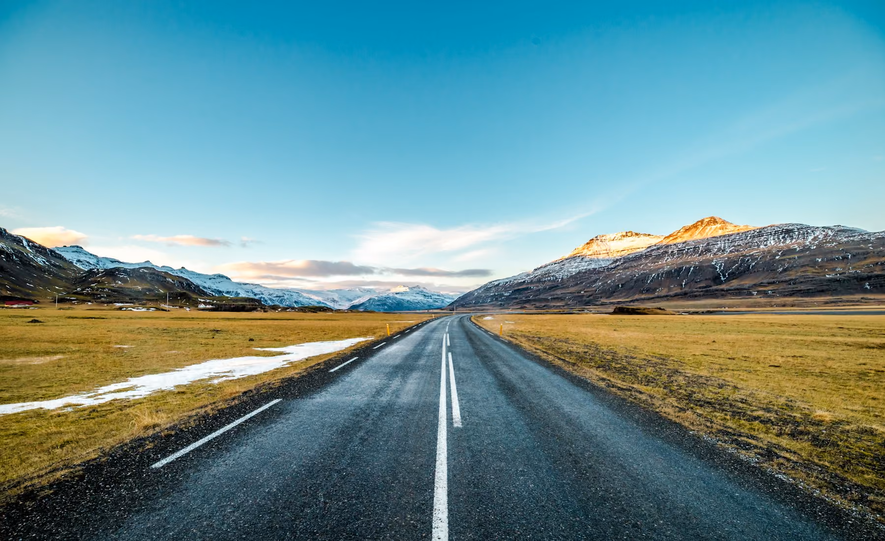An empty Icelandic road leading towards snow-dusted mountains in soft evening light