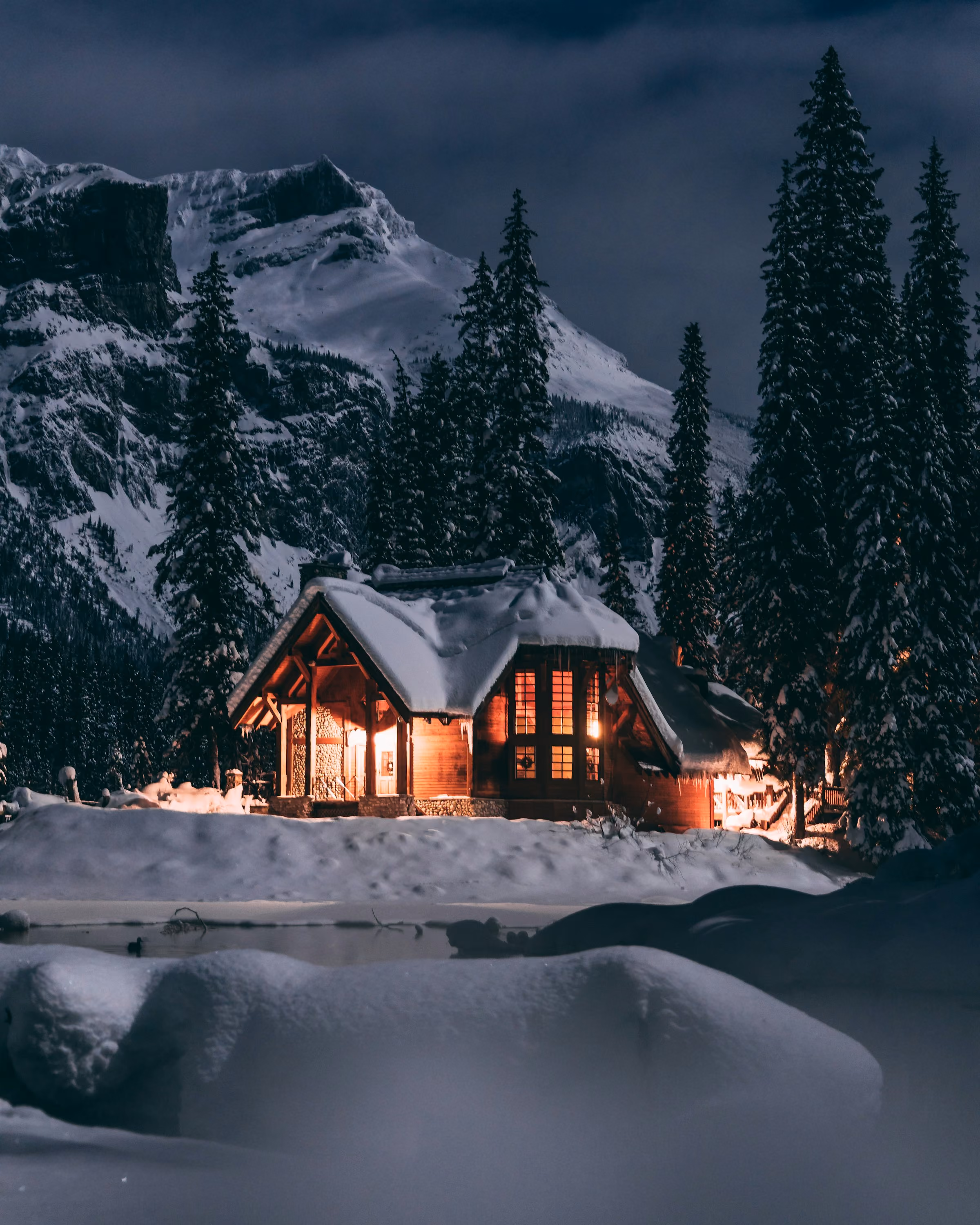 A lit wooden house among snowy pines below mountains