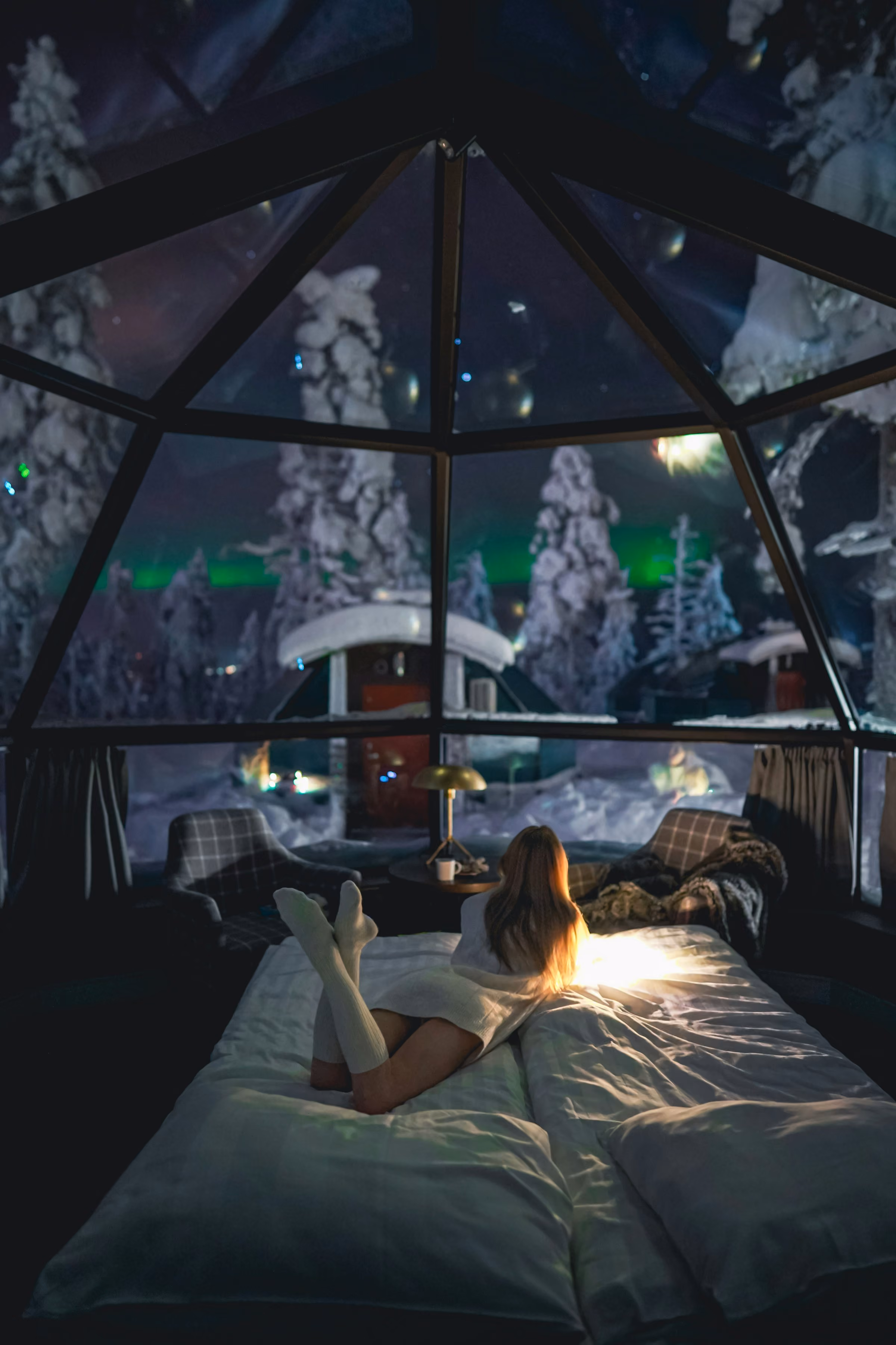 Looking up from a bed in a glass igloo at a green aurora above the trees