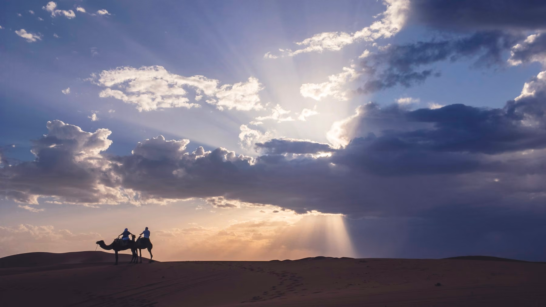 Two riders on camels silhouetted against sun rays and storm clouds at Erg Chebbi near Merzouga