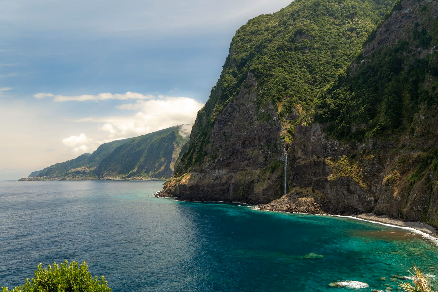 Green cliffs and a waterfall dropping towards a turquoise bay on the coast of Madeira