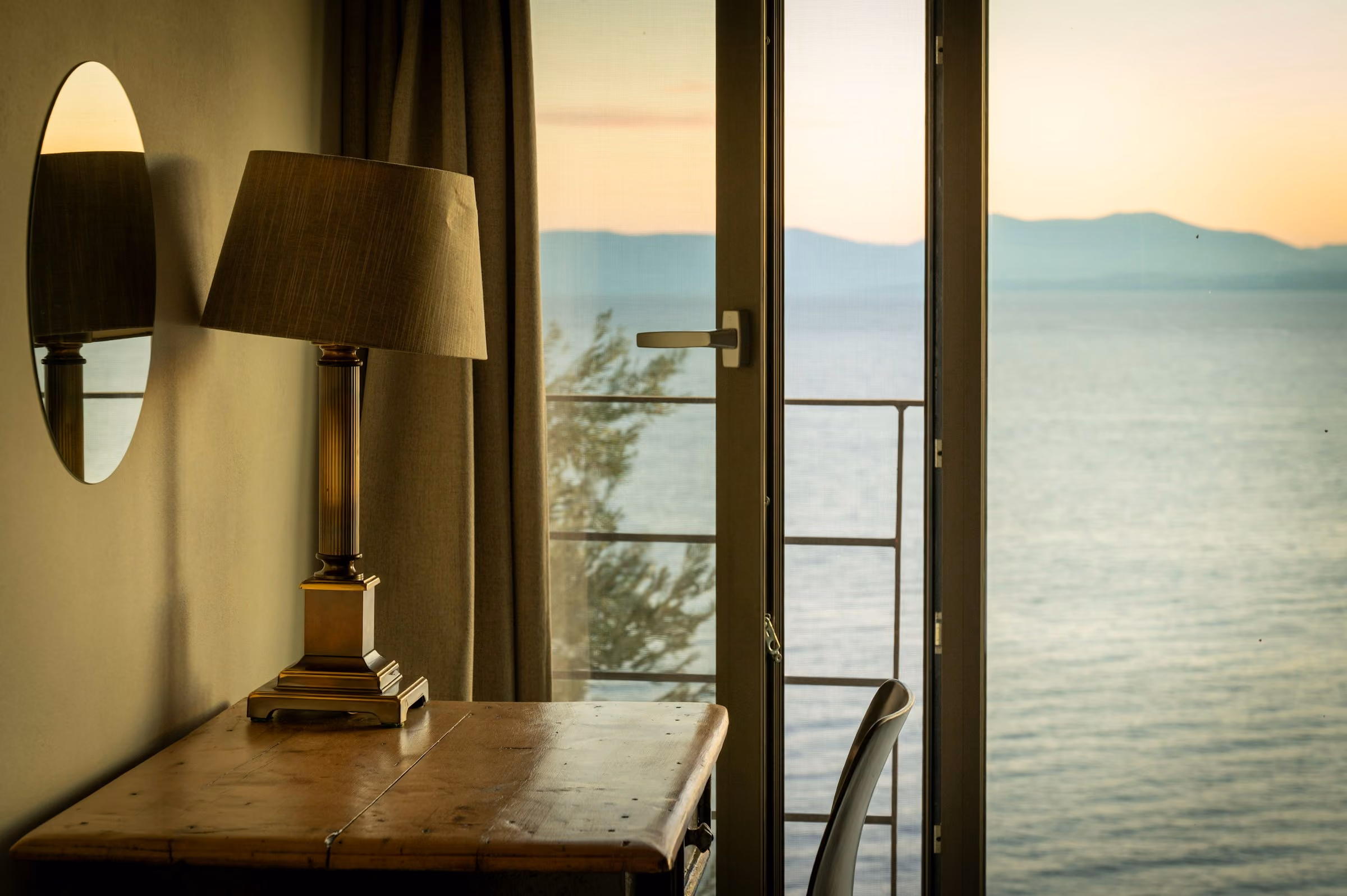 A brass lamp on a writing desk beside open balcony doors and a calm sea at sunset